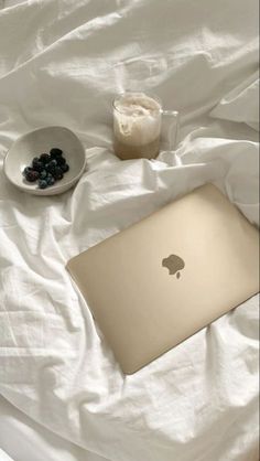 an apple laptop sitting on top of a bed next to a bowl of blueberries