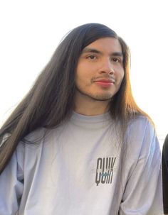 a man with long hair standing in front of a white wall wearing a gray shirt