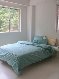 a bed with blue sheets and pillows in a white room next to two large windows