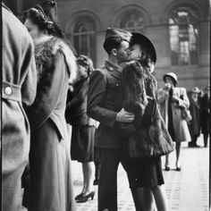 black and white photograph of two people kissing in front of a building with other people standing around