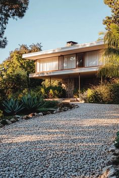 the house is surrounded by trees and rocks, with gravel on the ground in front