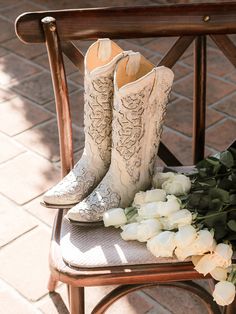 two pairs of cowboy boots sitting on top of a chair next to white roses and greenery