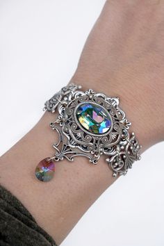 "Victorian crystal bracelet / Tudor bracelet  This detailed ornate filigree crystal bracelet is reminiscent of the elegance of the Victorian and Edwardian times Pair this crystal statement bracelet with dressy or casual outfits. A large sparkling glass crystal is the focal point of this romantic bracelet. The crystal color is called \"paradise shine\" and dances between hues of purple and green depending on the light. Bracelet secures at the back with a lobster clasp and extender chain for an adjustable fit. SIZE:  LENGTH: Bracelet will fit wrist sizes up to 7 inches. (If you need a larger size message me). WIDTH: measures 2 inches wide at the widest part.  * All metal components are of high quality silver plated brass * Nickel and lead free  * Presented in a black gift box with pillow- pe Fantasy Bracelet Art, Victorian Style Silver Crystal Jewelry, Victorian Silver Crystal Jewelry, Victorian Metal Bracelets With Jewels, Ornate Metal Bracelets With Jewels, Ornate Jeweled Bracelets As Gift, Ornate Jeweled Bracelets For Gifts, Ornate Silver Jeweled Bracelets, Jeweled Metal Crystal Bracelet As Gift