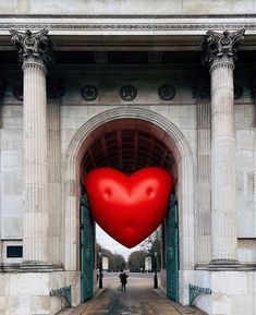 a large red heart shaped balloon floating in the air between two white pillars with people walking underneath it