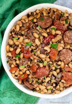 a white bowl filled with beans, sausage and other food on top of a green cloth