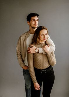 a man and woman standing next to each other in front of a gray background with their arms around each other