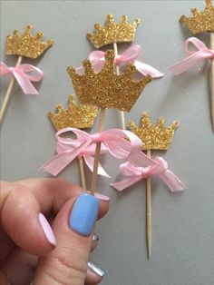 a hand holding some pink and gold cupcake toppers with bows on sticks in the shape of crowns