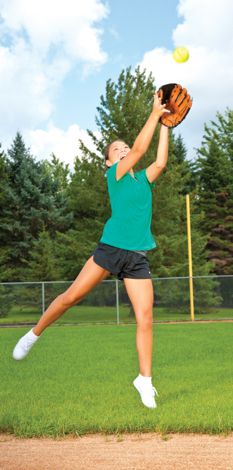 a woman catching a yellow ball in the air on top of a baseball field with trees in the background