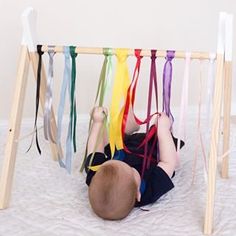 a baby laying on its back in front of a wooden frame with ribbons hanging from it