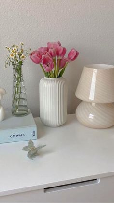 a white vase with pink flowers on top of a table next to a book and lamp