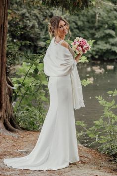 a woman in a white dress holding a bouquet and standing next to a river wearing a shawl