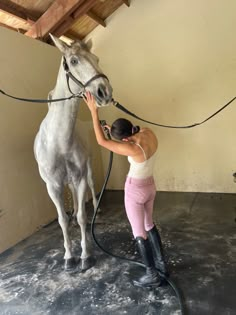 a woman is washing a horse with a hose in an indoor area that has concrete flooring