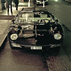 an old black sports car parked on the side of the road at night with people walking by