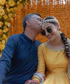 a man and woman sitting next to each other in front of an orange wall with flowers