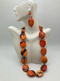 This piece displays a vibrant jewelry set consisting of a 20" necklace and matching earrings, both made from dyed turquoise. The necklace features large, flat oval beads with intense, vivid colors of orange and red, marbled with black and brown veining. These bold beads are spaced with small colored accents, adding a subtle contrast that enhances the overall vibrancy. The earrings complement the necklace with a matching oval bead, accompanied by small, stacked turquoise chips that mirror the necklace's color scheme. The bright and dynamic hues of this set create a striking and energetic look, perfect for making a bold fashion statement. The necklace features a post and hoop closure. Vibrant Jewelry, Handmade Orange Multi-strand Necklace, Orange Multi-strand Polished Beads Jewelry, Artisan Multi-strand Red Necklaces, Orange Amulet-style Jewelry With Large Beads, Oval Beads, Artisan Hand-strung Orange Beads Gems And Cabochons, Bold Fashion, Matching Earrings