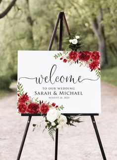 a welcome sign with red and white flowers hanging from it's easel in front of trees