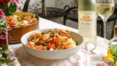 a white bowl filled with pasta and vegetables next to a bottle of wine on a table