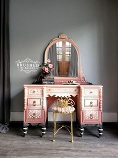 a pink vanity with mirror and stool in front of a gray wall, next to a window