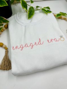 a white t - shirt with the word engaged now embroidered on it next to a plant