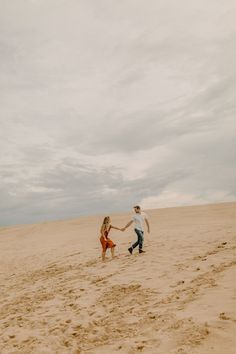 a man and woman holding hands while walking across a sandy beach on a cloudy day