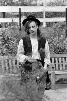 black and white photograph of a woman standing in front of a bench wearing a hat