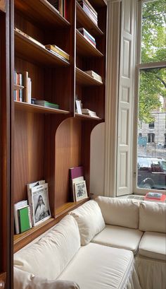 a white couch sitting in front of a window next to a wooden book shelf filled with books