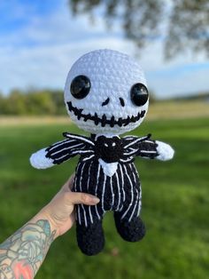 a hand holding a small knitted skeleton doll in a grassy field with trees and blue sky in the background