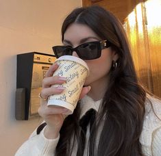 a woman wearing sunglasses drinking from a coffee cup in front of a wall mounted mailbox