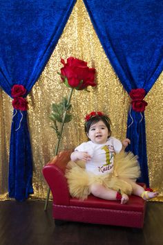 a baby sitting on a red chair in front of a blue curtain and a rose