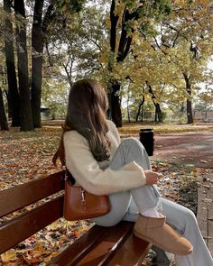 a woman sitting on top of a wooden bench in a park filled with fall leaves