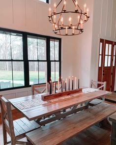 a wooden table with candles on it in front of large windows and a chandelier hanging from the ceiling