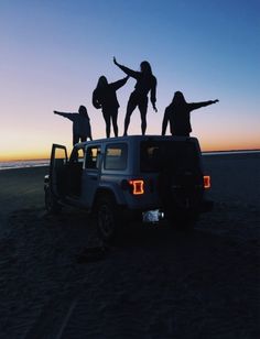 four people standing on the back of a jeep with their arms in the air at sunset