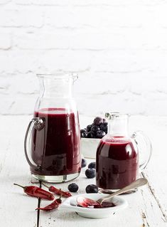 two pitchers of blueberries and raspberry juice on a white table with spoons