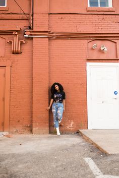 Black model with natural hair in purple rain tee, ripped denim, and white boots poses with back against red brick wall, one foot up on wall, and head tilted for vintage inspired photo session Urban Photo Shoot Outfits, Skyscraper Photoshoot, Merch Shoot, Downtown Dress, Urban Shoot, Pictures Downtown, Brand Shoots, Downtown Photoshoot, Urban Photoshoot