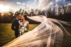 a bride and groom kissing in front of the sun at their outdoor wedding venue with veil blowing in the wind