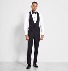 a man in a black tuxedo and bow tie stands against a white wall