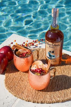 two copper mugs filled with drinks next to an empty bottle and some apples near the pool