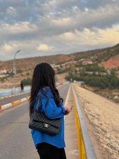 a woman standing on the side of a road looking at her cell phone and purse