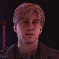 a man with blonde hair wearing a black shirt and looking at the camera while standing in front of a dark background