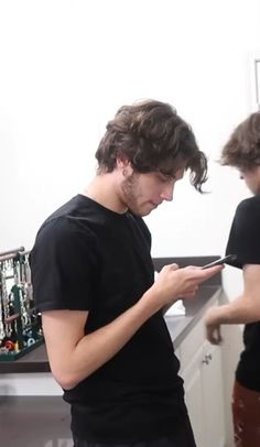 two young men standing in a kitchen looking at their cell phones and writing on paper