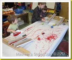 two young boys are painting with red paint on a large white table in a classroom