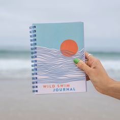 a hand holding a spiral notebook with the words wild swim journal written in front of it