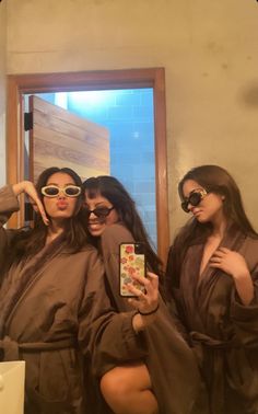 three women in brown robes and sunglasses taking a selfie with their cell phone while standing in front of a mirror