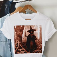 This adult unisex short sleeve tee features a dark and spooky tinted image of an afro witch in the woods. Crafted with soft cotton and a quality print, this tee will become your well-loved fall favorite. It's a great top to celebrate the spirit of Halloween, but the illustration works year round as well. Its ribbed knit collars, taping on the shoulders, and dual side seams ensure a better fit over time and maintain the garment's shape. Made with 100% cotton, this tee offers a comfortable retail fit and light fabric weighing 4.2 oz/yd² (142 g/m²). With its tear away label and true-to-size fit, our witchy tee is perfect for those who want style, comfort, and a magical design. Available in sizes XS-5XL. Easily customizable upon request. Shop The Trini Gee for more: https://www.etsy.com/shop/t Fall Cotton T-shirt With Character Print, Fitted Band Merch T-shirt For Fall, Unisex Graphic Print T-shirt For Fall, Unisex Halloween Graphic Print Top, Unisex Graphic Print Top For Halloween, Cotton Character Print T-shirt For Fall, Cotton T-shirt With Character Print For Fall, Unisex Fall Short Sleeve T-shirt, Unisex Short Sleeve T-shirt For Fall