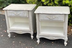 two white nightstands sitting next to each other in front of some bushes and trees