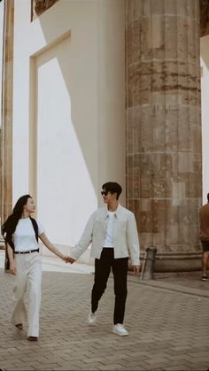 a man and woman holding hands while walking down a brick walkway in front of columns