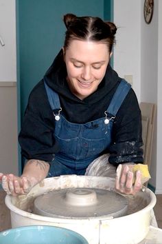 a woman in overalls is making something out of clay with her hands and smiling at the camera