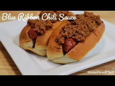 two hotdogs with chili on them sitting on a white square plate that says blue ribbon chili sauce