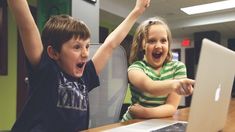 two young children sitting at a desk with their arms in the air and looking at a laptop