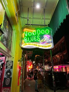 the neon sign for voodoo blues is lit up in green and yellow colors, with people walking down the street below